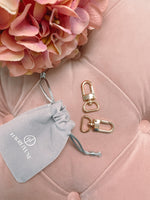 Two gold Luggage Tag Clip pieces beside a drawstring pouch on a pink cushioned surface, tagline Your Handbags Forever Starts Here.